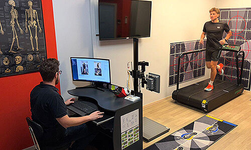 Übersicht eines Analyseraums mit Laufband, Monitoren und Fachpersonal, das die Laufbewegung eines Sportlers auswertet